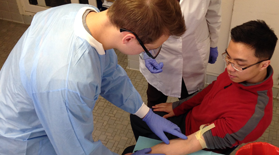 A medical student in a surgical gown practices locating a vein on a peer's arm during a blood draw training session.